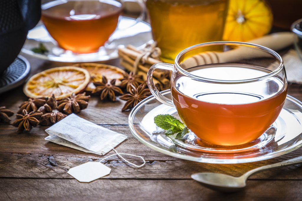 Uma xícara transparente com chá sobre uma mesa, onde também estão um sachê de chá, uma colher, anis estrelados, canela em casca e rodelas de laranja